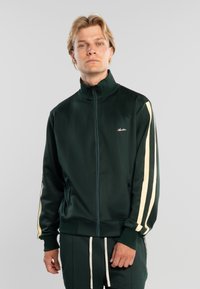 Hombre rubio con chaqueta verde oscuro con cremallera y franjas crema en las mangas, y pantalones a juego con cordones crema, de pie frente a un fondo gris.