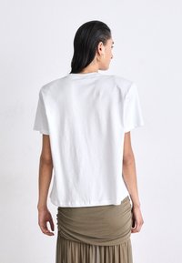T-shirt bianca in cotone con maniche corte, taglio ampio e colletto rotondo, mostrata di schiena. Caratterizzata da una superficie liscia e un design minimale.