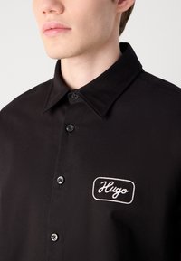 Zwarte overhemden met een kraag, met een geborduurd wit "Hugo" logo op een rechthoekig patch en zwarte knopen aan de voorkant.