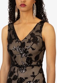 Schwarzes florales Spitzenkleid mit V-Ausschnitt, verziert mit Pailletten. Auffällige silberne Perlenohrringe ergänzen den Look.