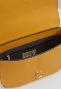 Žlutá kožená taška přes tělo s hladkým povrchem, vybavená zipovou vnitřní kapsou a zlatými kovovými detaily.