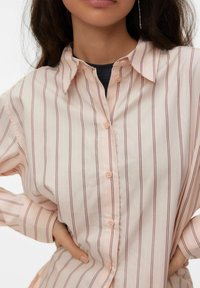 Femme portant une chemise rose clair à boutons avec des rayures verticales foncées, les mains sur les hanches, et de longs cheveux foncés partiellement visibles.