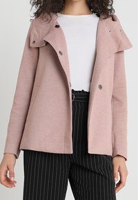 Rosa, strukturerad jacka med huva, som har en enkel knäppning och två sidofickor. Buren över en vit topp och svarta randiga byxor.