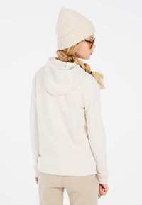 Felpa beige con cappuccio regolabile, morbida al tatto, e tasca a marsupio, abbinata a un berretto di maglia cremoso e testurizzato e occhiali da sole neri.