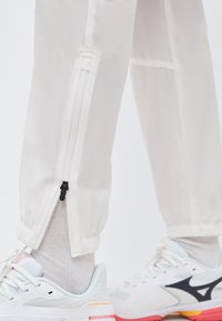 Φερμουάρ στα μανίκια λευκών αθλητικών παντελονιών φόρμας, φορεμένα με λευκά αθλητικά παπούτσια και ανοιχτόχρωμες γκρι κάλτσες, εμφανιζόμενα σε όρθιο άτομο.