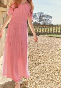 Maxiklänning i ljusrosa, tillverkad av flödande tyg. Har avskurna ärmar, veck och en avslappnad design. Bärs med vita skor.
