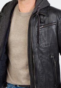 Zwarte leren jas met een grijze capuchon, voorzien van ritszakken, gedetailleerde stiksels en een zachte beige trui eronder.