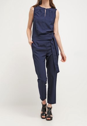 Femme portant une combinaison sans manches bleu marine avec ceinture à la taille, détail en forme de goutte d'eau à l'avant, et sandales noires à talons ouverts, debout devant un fond uni.