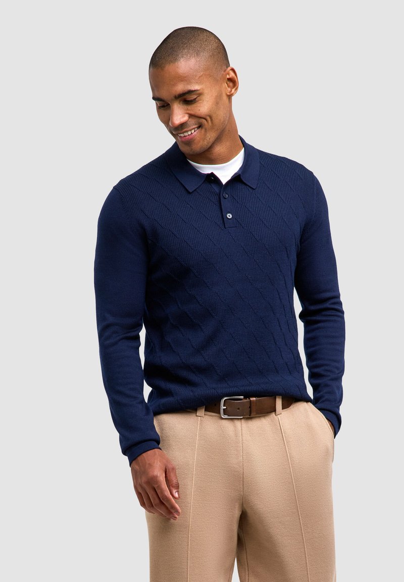 Polo sweater a maniche lunghe di colore blu navy con una texture a diamante, dotato di colletto e chiusura a tre bottoni, abbinato a pantaloni beige su misura.