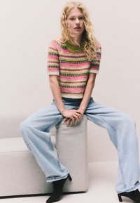 Rövid ujjú, csíkos pulóver rózsaszín, zöld és krémszínben; világoskék, bőszárú farmerral és fekete magassarkú csizmával párosítva, világosszürke tömbön ülve.