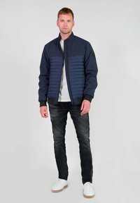 Marine bomberjack met gevoerde voorkant en geribde manchetten, gecombineerd met donkere jeans en witte sneakers. Gladde stof, ritssluiting en logo detail.