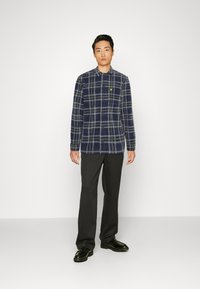 Camicia a quadri in blu navy e grigio con chiusura a bottoni, maniche lunghe e logo a patch. Abbinata a pantaloni scuri e scarpe nere.