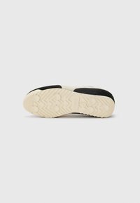 Suela de zapatilla negra y crema con una superficie de goma texturizada, que presenta patrones de agarre circulares y un diseño de banda de rodadura en zigzag para mejorar la tracción.