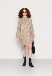 Vestido de malha em bege com padrões texturizados, decote alto e mangas balão. Usado com botins pretos e uma mala preta.