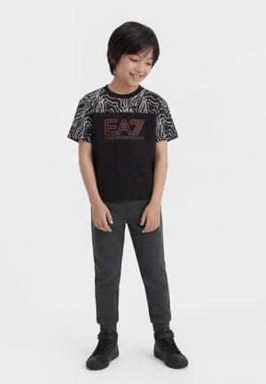 Boy wearing black and gray EA7 Emporio Armani t-shirt with patterned sleeves, dark jogger pants, and black high-top sneakers, standing and smiling.