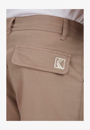 Bežīgas kokvilnas bikses ar aizmugurējo kabatu, kurā ir balts izšūts logo. Materiāls ir gluds un tam ir maiga spīduma.