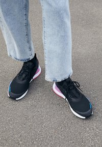 Black athletic sneakers with mesh upper, glossy accents, and colorful sole in pink and white, paired with cuffed light denim jeans.