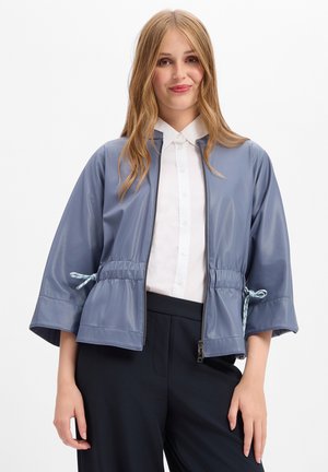 Young woman with long blonde hair wearing a blue zip-up jacket over a white buttoned shirt and black pants against a plain white background.