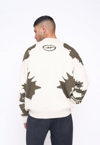 Trui in off-white met een olijfgroen abstract patroon. Heeft een ronde hals, lange mouwen en een geribbelde zoom. Logo detail op de achterkant aanwezig.
