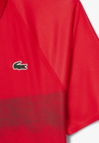 Röd polo-skjorta med en texturerad gradient, korta ärmar, och en grön krokodil-logotyp på bröstet. Tillverkad av ett mjukt tyg.