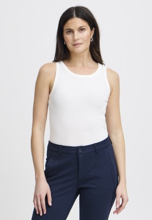 Femme aux longs cheveux foncés portant un haut blanc sans manches à côtes et un pantalon bleu marine, debout devant un fond clair uni.