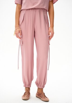 Kobieta ubrana w różowe cargo z elastycznymi mankietami i pasujące różowe sznurowane buty, stojąca na białym tle.