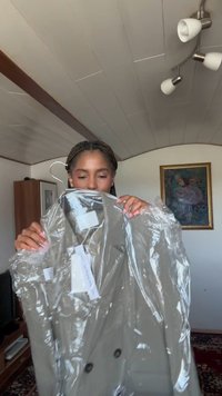 Beige double-breasted trench coat in clear plastic, featuring large black buttons and tags attached, held by a person in a room.