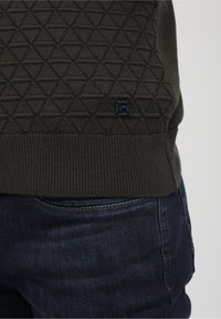 Pull en maille vert foncé présentant un motif triangulaire texturé, une bordure côtelée et une petite étiquette logo bleue près du bas. Porté avec un jean bleu.