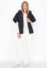 Marineblauw gewatteerd hooded vest met een rits aan de voorkant en korte mouwen, gecombineerd met witte wijde broeken en lichte sneakers.