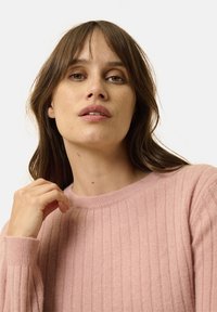 Suéter de punto rosa con cuello redondo, mangas largas y textura suave; la modelo sostiene suavemente su mano cerca del escote.