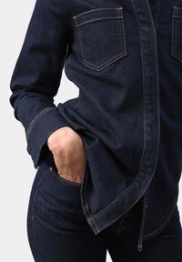 Mørk denimskjorte med buet kant, knaplukning, to brystlommer og kontrastfarvet orange syninger. Kombineret med matchende denimbukser.