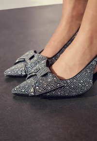 Čierne baleríny s ostrou špičkou, pokryté striebornými kamienkami, so veľkou mašľou na priehlavku a hladkým textúrou.