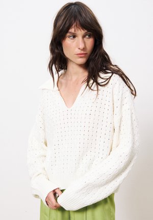 Pull en tricot blanc avec un col V et un col, présentant un motif de tricot ouvert, des manches longues et des poignets côtelés. Assorti à un pantalon vert.