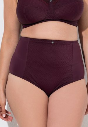 Femme portant un sous-vêtement texturé violet foncé taille haute et un soutien-gorge assorti, debout devant un fond uni.