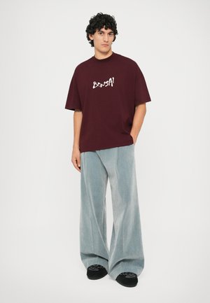 T-shirt oversized em cor burgundy com "BONSAI" em letras brancas, combinado com calças de corduroy de perna larga em azul claro. Sapatos pretos com detalhes decorativos.