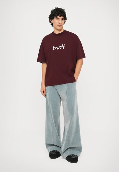 Burgunderfarbene oversized T-Shirt mit dem Schriftzug "BONSAI" in weißen Buchstaben, kombiniert mit hellblauen weit geschnittenen Cordhosen. Schwarze Schuhe mit dekorativen Akzenten.