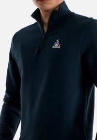 Navy blue quarter-zip pullover in soft fabric with a logo on the left chest. Features long sleeves and a high collar for added warmth.