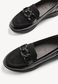 Paire de mocassins noirs avec dessus en daim, bordure en cuir verni et grandes décorations brillantes en forme de maillons de chaîne sur le devant.