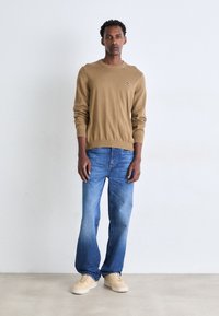 Hnedý pletený sveter s okrúhlym výstrihom, skombinovaný s pohodlnými modrými džínsami a béžovými teniskami. Obsahuje malé logo na svetri.