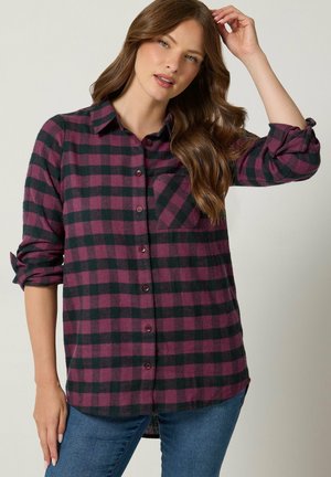 Mujer con cabello largo y castaño que lleva una camisa de cuadros morada y negra abotonada y vaqueros azules, ajustándose el cabello con una mano.