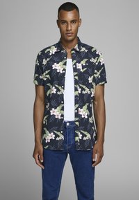 Camisa de flores de manga corta en navy con hojas verdes y flores blancas, que presenta un diseño de botones y una camiseta interior blanca lisa debajo.