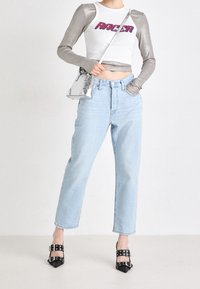 Person bär en vit croppad topp med texten "RACER" och glänsande långa ärmar, ljusblå högmidjade jeans, svarta klackskor med spänne och en silverfärgad axelväska.