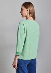 Ljusgrön stickad tröja med trekvartsärmar, med en ribbad halsringning och mudd, i kombination med blå denim-jeans.