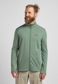 Veste zippée verte avec un motif texturé, col montant et deux poches latérales. Le tissu semble léger et respirant. Logo visible sur la poitrine.