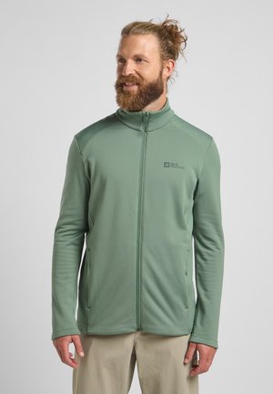 Groene ritsjack met een getextureerd patroon, hoge kraag en twee zijzakken. De stof lijkt lichtgewicht en ademend. Logo zichtbaar op de borst.