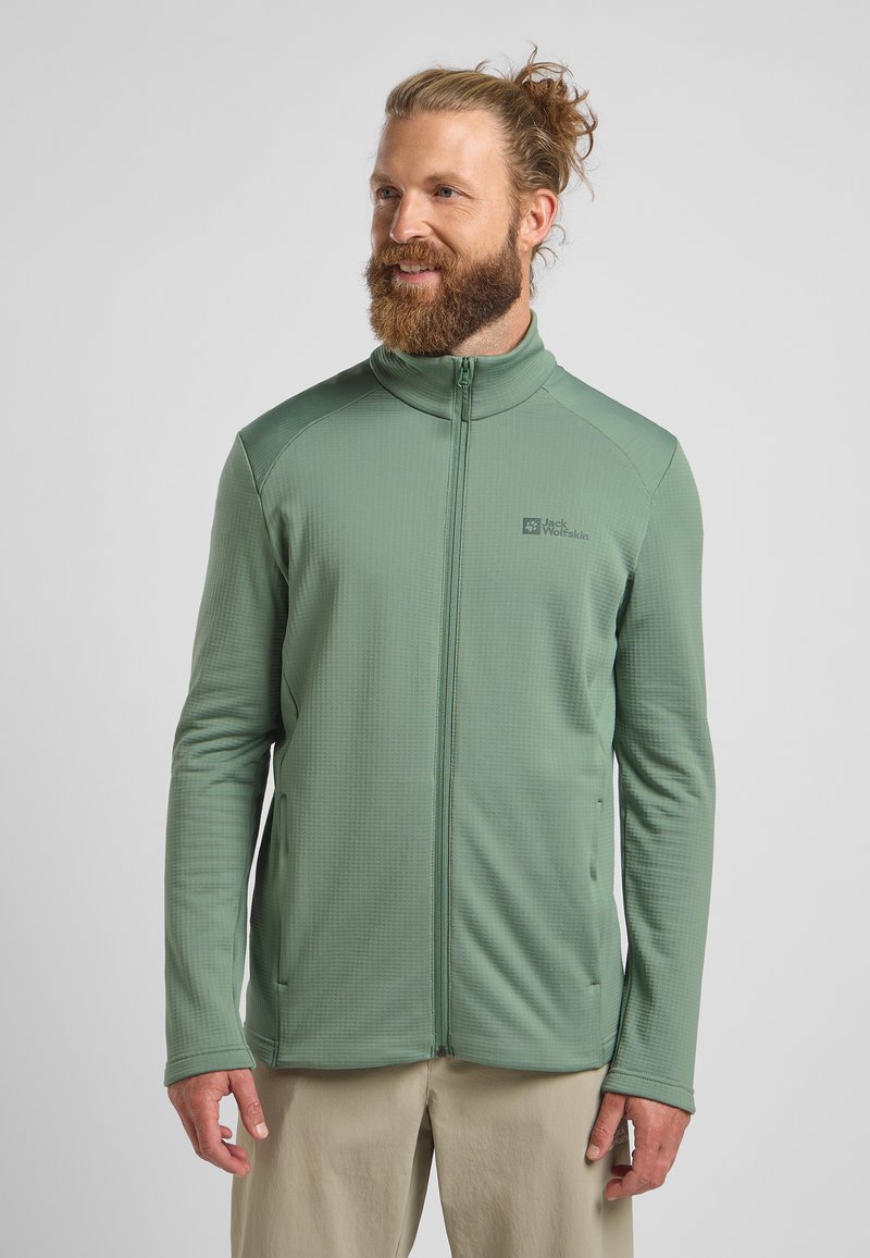 Veste zippée verte avec un motif texturé, col montant et deux poches latérales. Le tissu semble léger et respirant. Logo visible sur la poitrine.