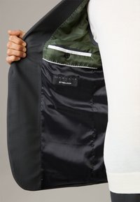 Blazer da uomo con un tessuto esterno grigio scuro e strutturato. Foderato con materiali lisci neri e verdi, presenta una tasca con bottone e etichetta.