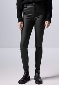 Hoge taille, nauwsluitende zwarte neplederen broek met glanzende textuur, zijzakken en riemlussen, gecombineerd met zwarte enkellaarsjes.