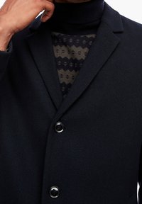 Marineblå vævet frakke med skrå krave, to knapper og tekstureret stof. Underneden er en mønstret mørk og grøn sweater delvist synlig.