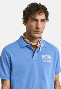 Ljusblå pikétröja med strukturtyg; har en krage och en vit "SEASIDE GANT RESORT" logotyp. En mönstrad scarf bärs runt halsen.